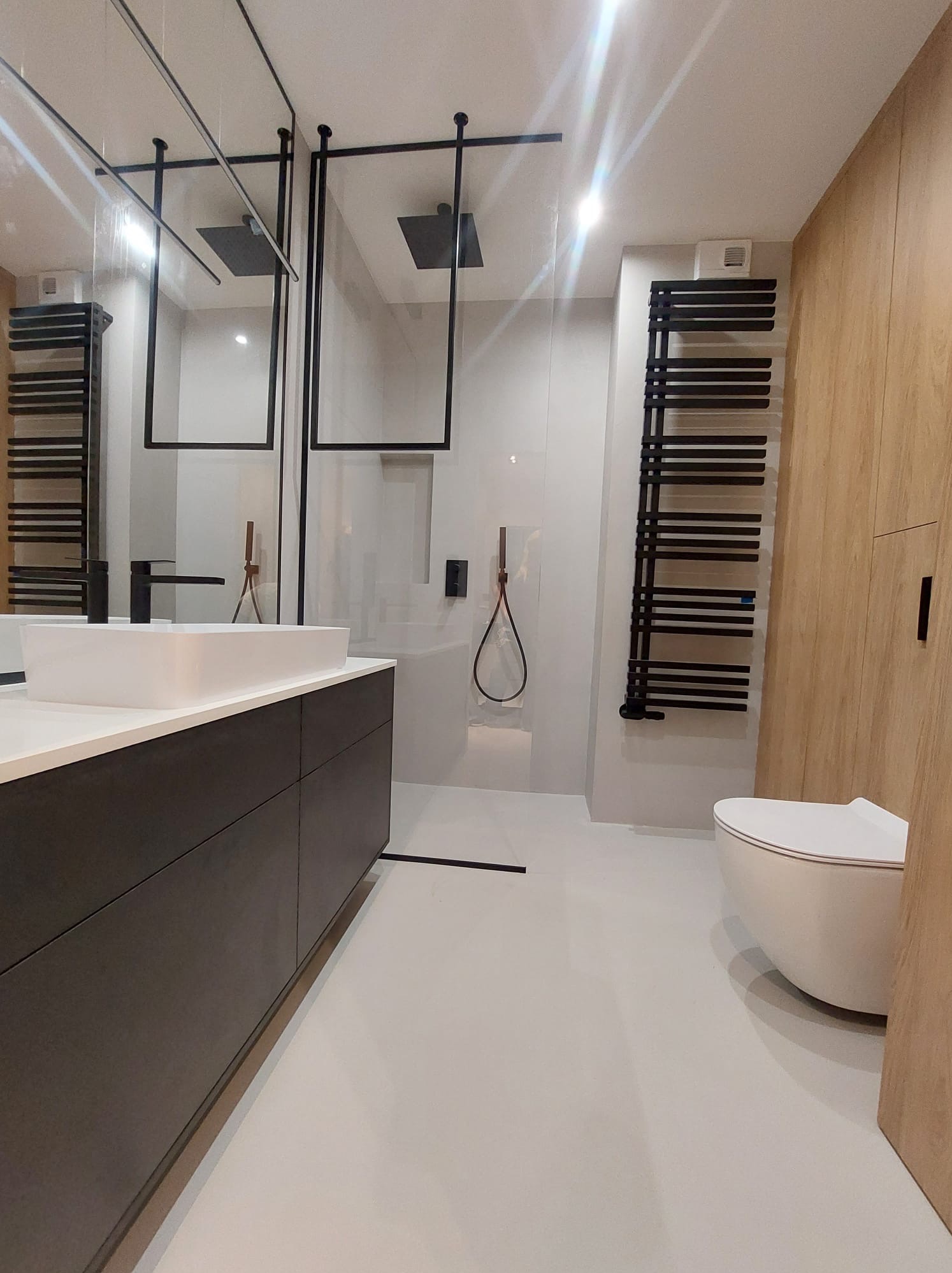 Nowoczesna łazienka z białą posadzką żywiczną, kabiną prysznicową typu walk-in z czarnymi ramami, umywalką nablatową i grzejnikiem drabinkowym.