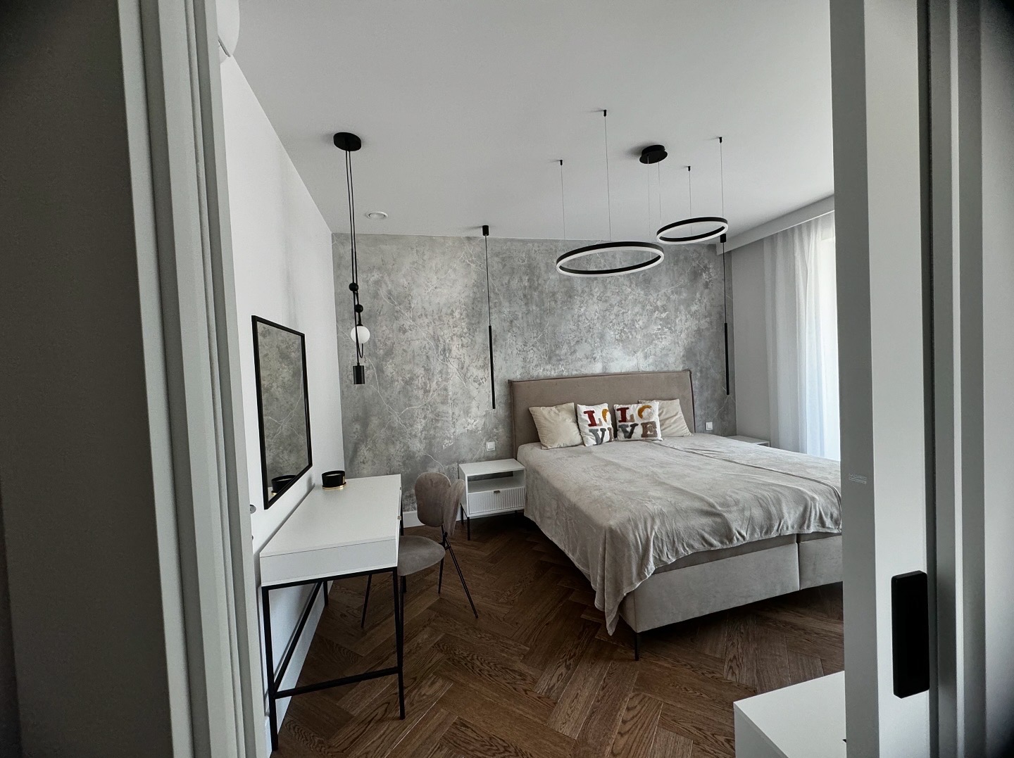 Nowoczesna sypialnia z parkietem w jodełkę, szarą ścianą dekoracyjną, łóżkiem z poduszkami z napisem i wiszącymi lampami LED w kształcie okręgów.