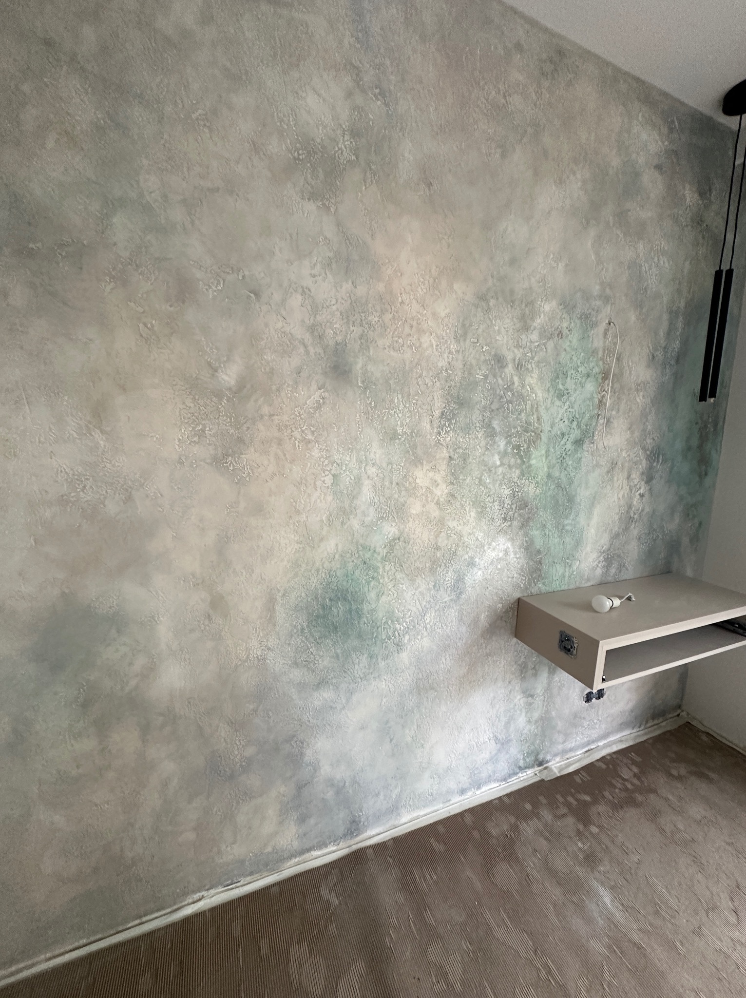 Ściana z dekoracyjną, nieregularną fakturą w odcieniach szarości i zieleni, widoczna półka z żarówką i fragment podłogi z brązowymi panelami.