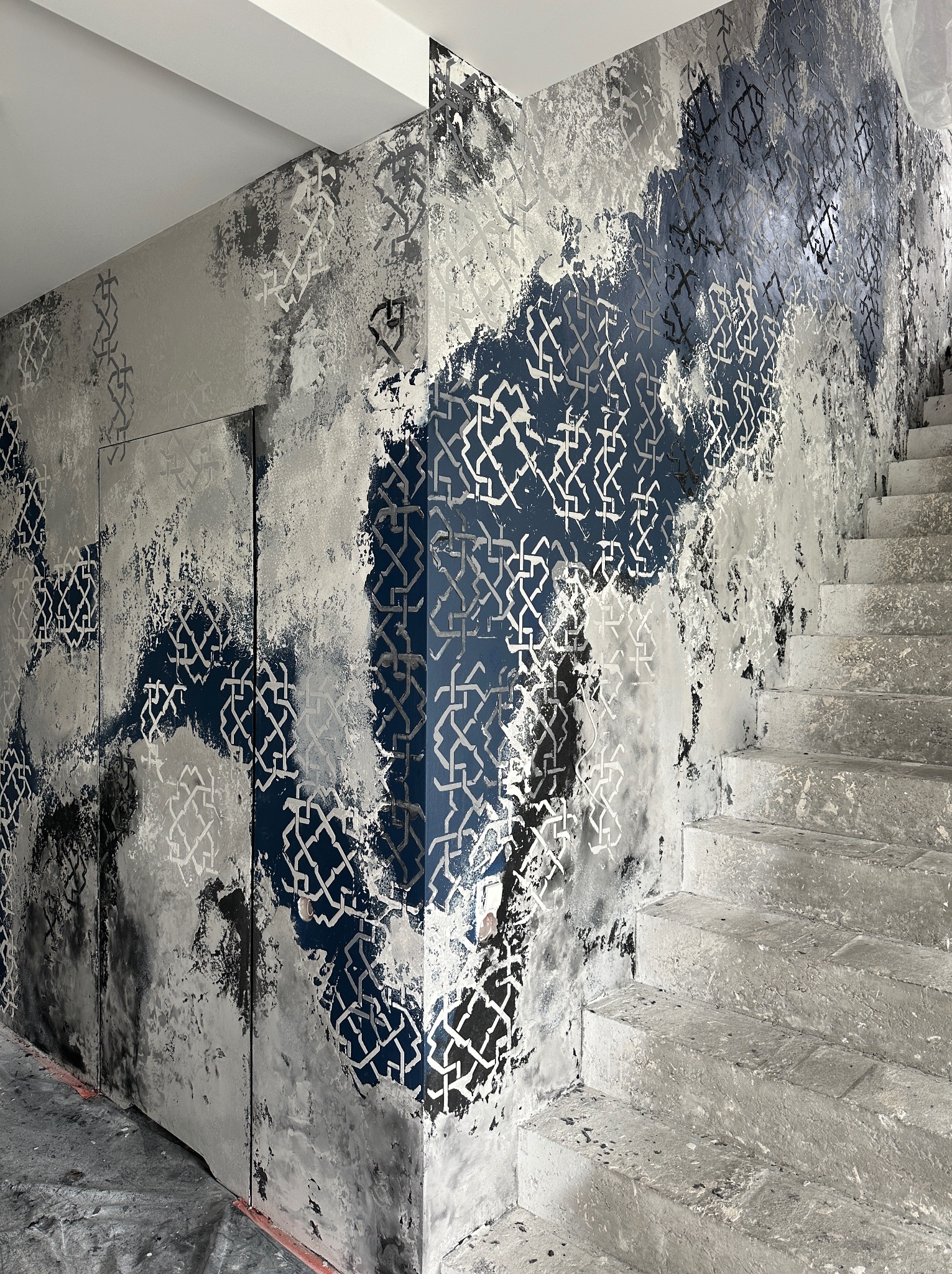 Ściana z dekoracyjną fakturą w odcieniach szarości i granatu, wzór geometryczny, obok schody betonowe w surowym stanie.
