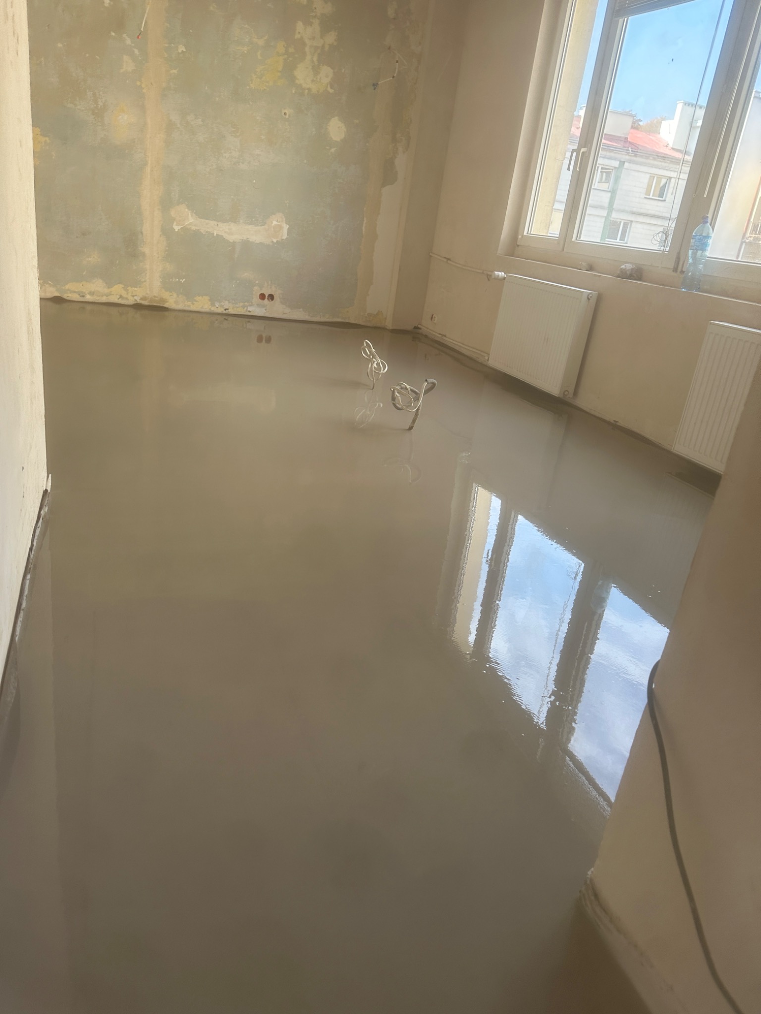 Świeżo wylana, gładka, samopoziomująca wylewka podłogowa w jasnym pomieszczeniu z oknem, widoczne mieszadła do zaprawy na posadzce, ściany przygotowane do dalszych prac wykończeniowych.