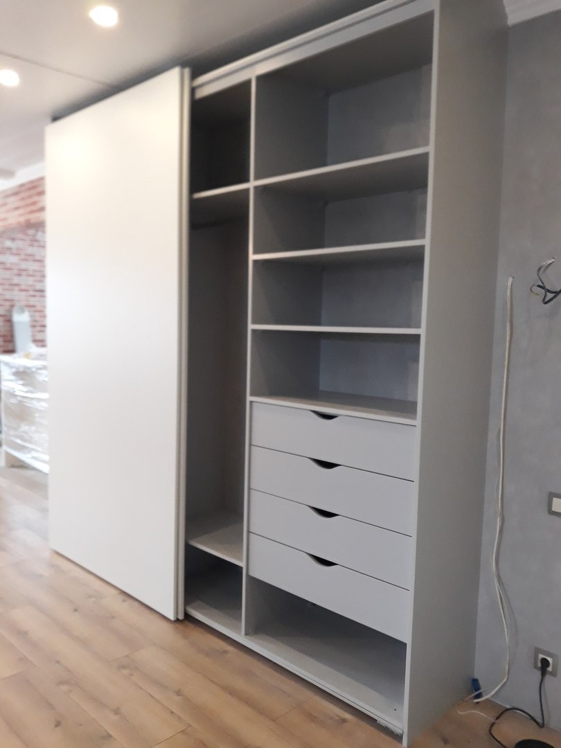 Szara szafa przesuwna z półkami i czterema szufladami, widoczna na drewnianej podłodze w pokoju z ceglaną ścianą w tle.