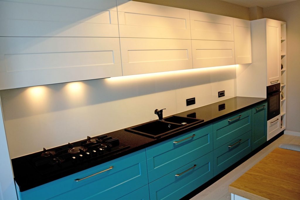 Nowoczesna kuchnia z turkusowymi szafkami dolnymi i białymi górnymi, czarny blat z wbudowanym zlewem i płytą gazową, oświetlenie LED pod szafkami górnymi.