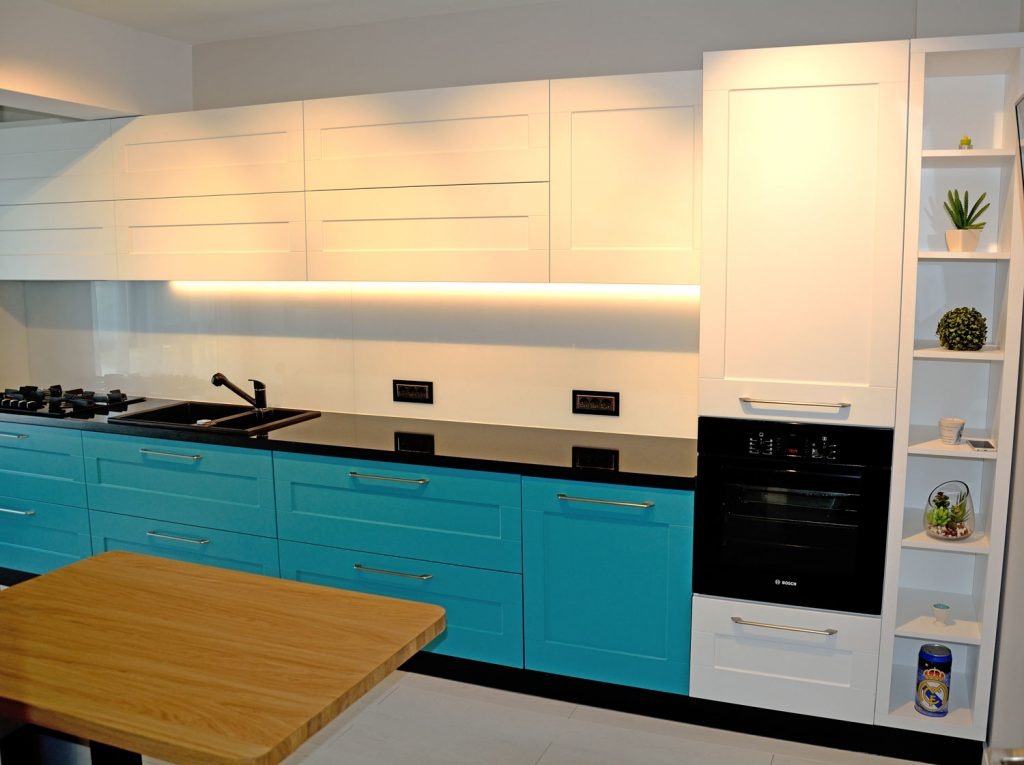 Nowoczesna kuchnia z turkusowymi szafkami dolnymi, białymi górnymi i czarnym blatem, oświetlona listwą LED, z widocznym fragmentem drewnianego stołu.