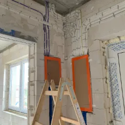 Wnętrze w trakcie prac wykończeniowych, widoczne rozprowadzone przewody elektryczne w białych i niebieskich peszlach, zamaskowane otwory w ścianie pomarańczową taśmą oraz drewniana drabina oparta...