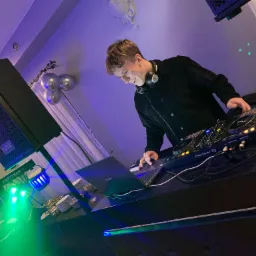 DJ w trakcie występu, miksuje muzykę na sprzęcie Pioneer DJ, obok laptop Asus, kolumna głośnikowa WAV i oświetlenie sceniczne BeamZ Partybar. W tle dekoracja z balonów.