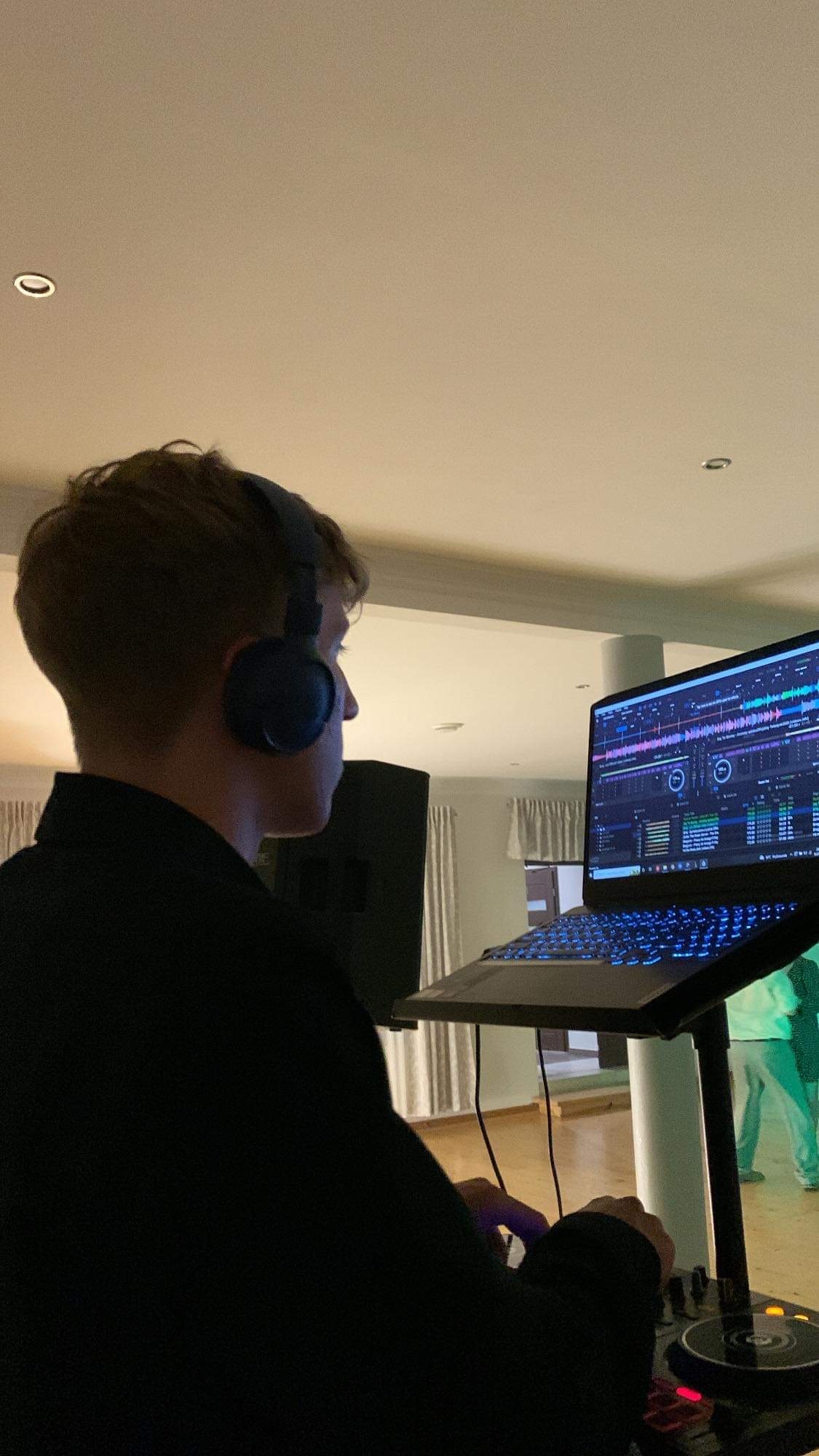 DJ w słuchawkach, z profilu, obsługujący konsoletę DJ-ską z laptopem oświetlonym niebieskim światłem, w tle sylwetki osób.