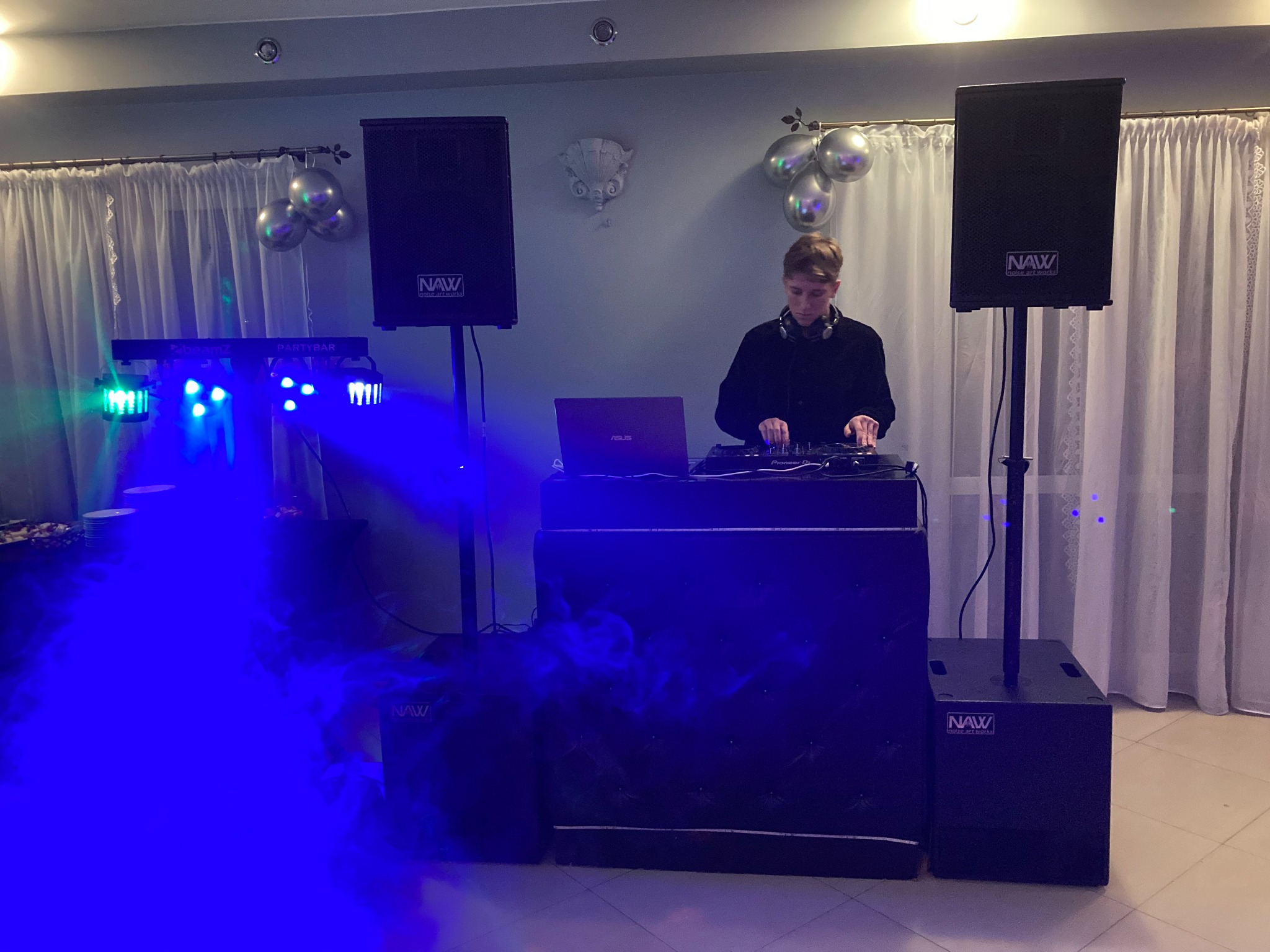 DJ w trakcie występu, miksujący muzykę na profesjonalnym sprzęcie Pioneer DJ, z laptopem Asus, nagłośnieniem marki NAW i efektami świetlnymi Partymuz, w tle dekoracja z balonów i białe zasłony.