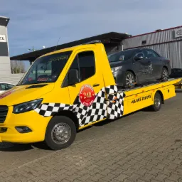 Ż&oacute;łty samoch&oacute;d pomocy drogowej z czarno-białą szachownicą na boku, transportujący uszkodzony szary samoch&oacute;d osobowy marki Toyota na tle salonu samochodowego Toyota Katowice.