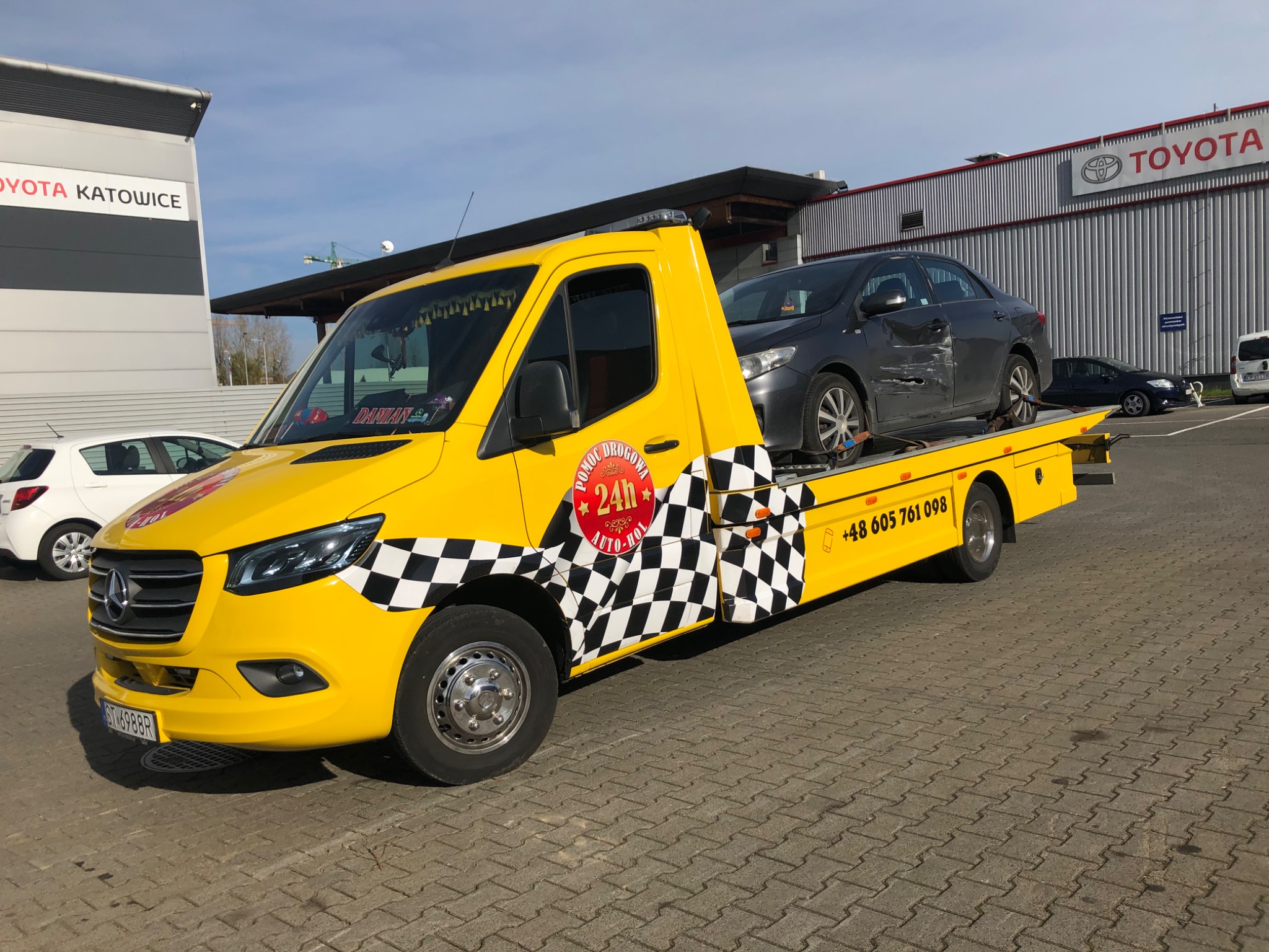 Żółty samochód pomocy drogowej z czarno-białą szachownicą na boku, transportujący uszkodzony szary samochód osobowy marki Toyota na tle salonu samochodowego Toyota Katowice.