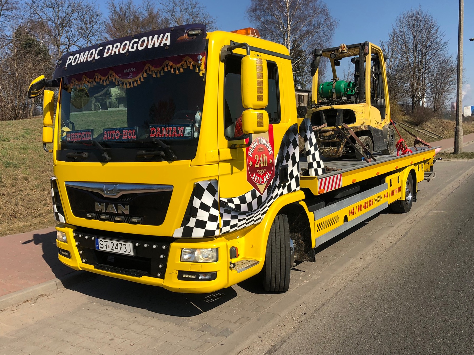 Żółta laweta z napisem 'Pomoc Drogowa', przewożąca uszkodzony wózek widłowy, z dekoracjami w szachownicę i logo '24h Auto-Hol'.
