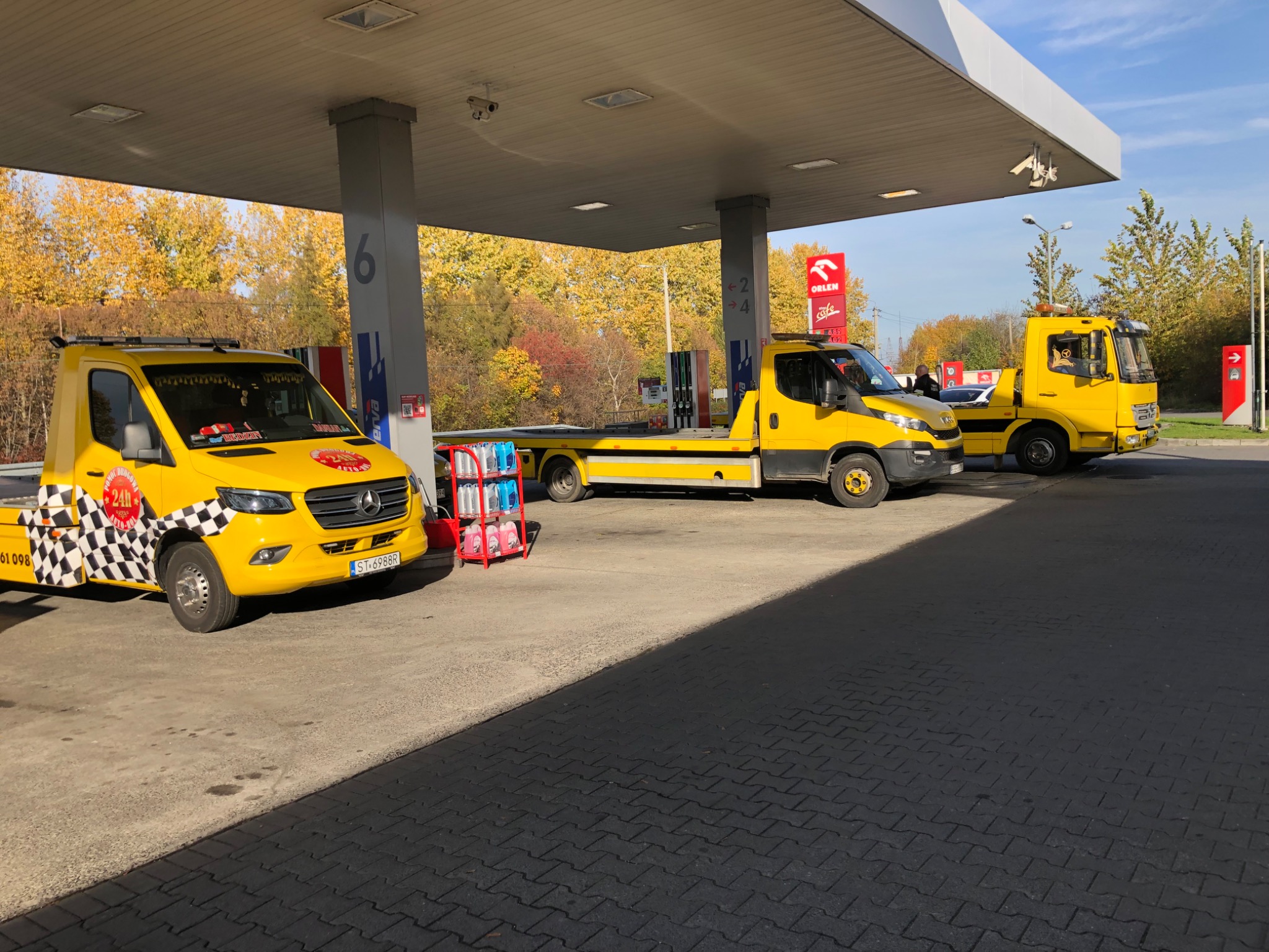 Trzy żółte pojazdy pomocy drogowej zaparkowane pod zadaszeniem stacji benzynowej Orlen w słoneczny dzień. Pierwszy pojazd z czarno-białą szachownicą i napisem 'Pomoc Drogowa 24h'.