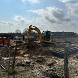 Only Trees - Marcin Miller - Żółta koparka Komatsu wykonuje wykop pod fundamenty na piaszczystym terenie budowy, widoczne szalunki i zbrojenia, w tle składowane bloczki Termoton, ogrodzenie i drzewa pod błękitnym niebem.