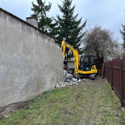Only Trees - Marcin Miller - Żółta koparka Komatsu częściowo wyburza szarą, betonową ścianę budynku na zewnątrz, z gruzem leżącym na trawie, pod pochmurnym niebem.