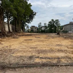 Przygotowany teren pod budowę z widocznymi śladami po maszynach, w tle budynki mieszkalne i drzewa, pochmurne niebo.