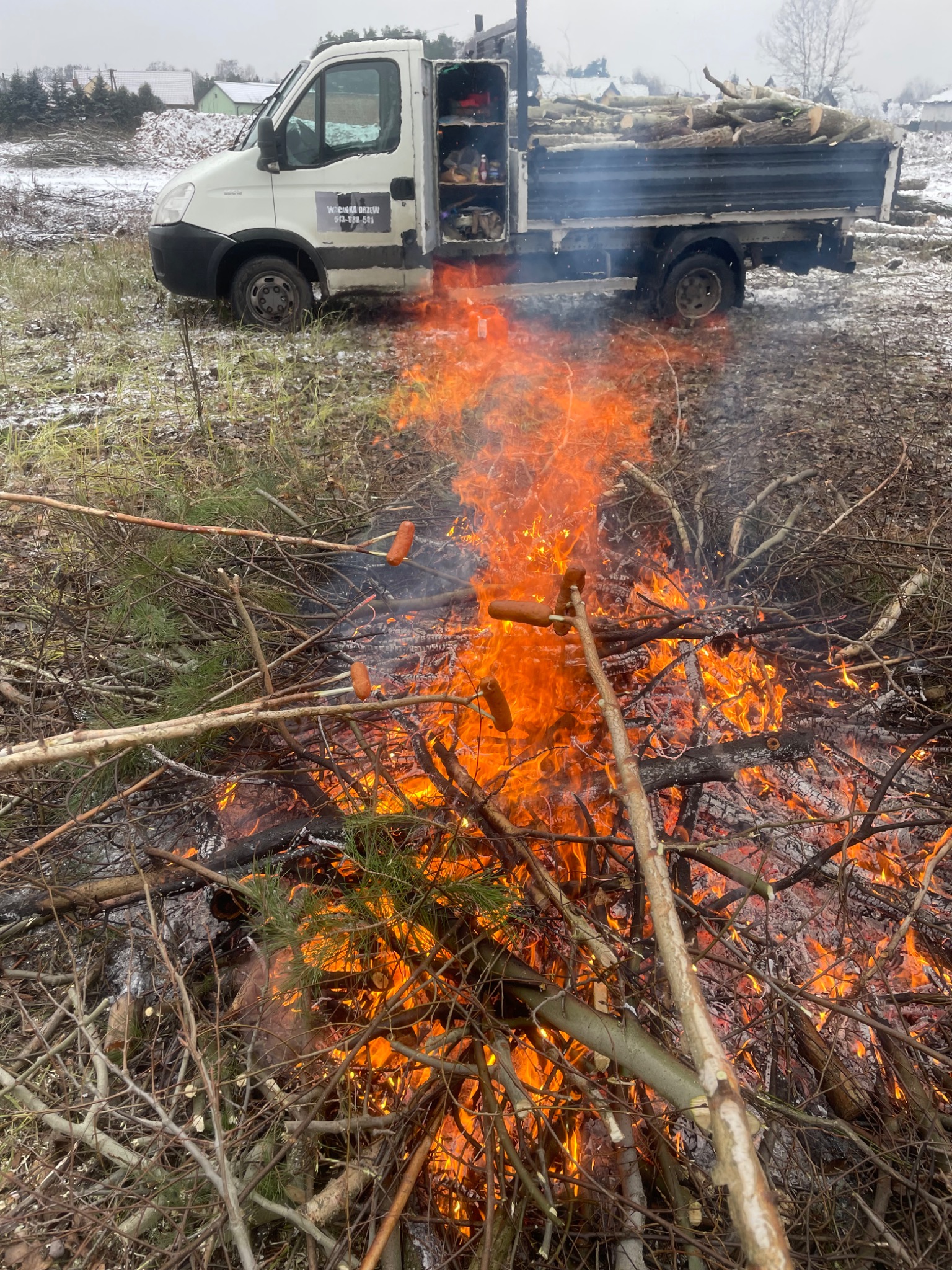 Ognisko z palonych gałęzi i piekących się kiełbasek, na tle samochodu dostawczego z napisem 'Wycinka Drzew', w zimowej scenerii.
