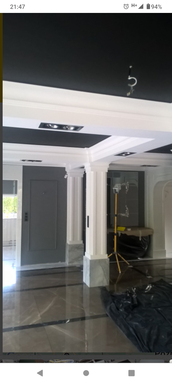 Eleganckie wnętrze z czarnym sufitem, białymi sztukateriami i kolumnami z marmurową bazą, widoczny fragment osłony podłogi i lampa budowlana.