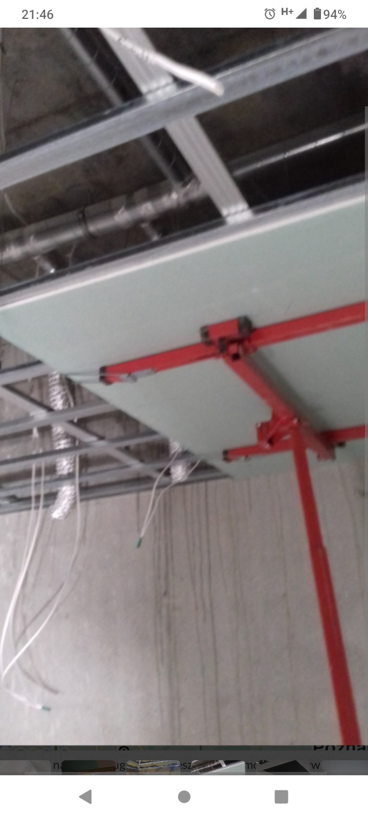 Montaż płyty gipsowo-kartonowej na metalowym ruszcie sufitowym przy użyciu czerwonego podnośnika. Widoczne przewody i rury pod sufitem.