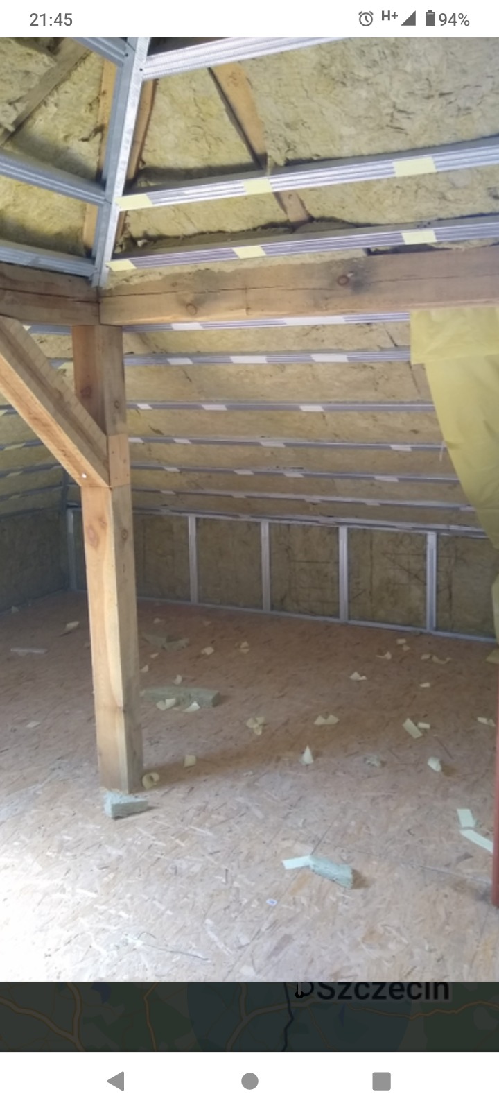 Poddasze w trakcie ocieplania wełną mineralną, widoczne drewniane elementy konstrukcyjne i metalowe profile, na podłodze leżą fragmenty izolacji.