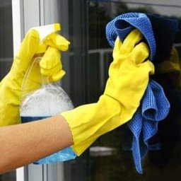 Pucus - Ręce w żółtych rękawicach spryskują szybę płynem z butelki, druga ręka trzyma niebieską ściereczkę z mikrofibry, odbicie w szybie.