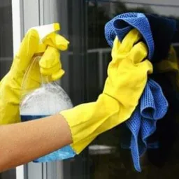 Ręce w żółtych rękawicach spryskują szybę płynem z butelki, druga ręka trzyma niebieską ściereczkę z mikrofibry, odbicie w szybie.