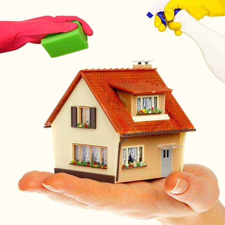 Miniaturowy domek z czerwoną dachówką trzymany na dłoni, czyszczony gąbką i spryskiwaczem z płynem, symbolizujący dbałość o czystość i higienę.