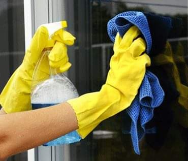 Ręce w żółtych rękawicach spryskują szybę płynem z butelki, druga ręka trzyma niebieską ściereczkę z mikrofibry, odbicie w szybie.