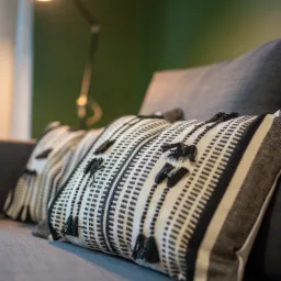 Dwa dekoracyjne poduszki w czarno-białe paski z frędzlami, ułożone na szarej sofie, w tle zielona ściana i lampka biurkowa dająca ciepłe światło.