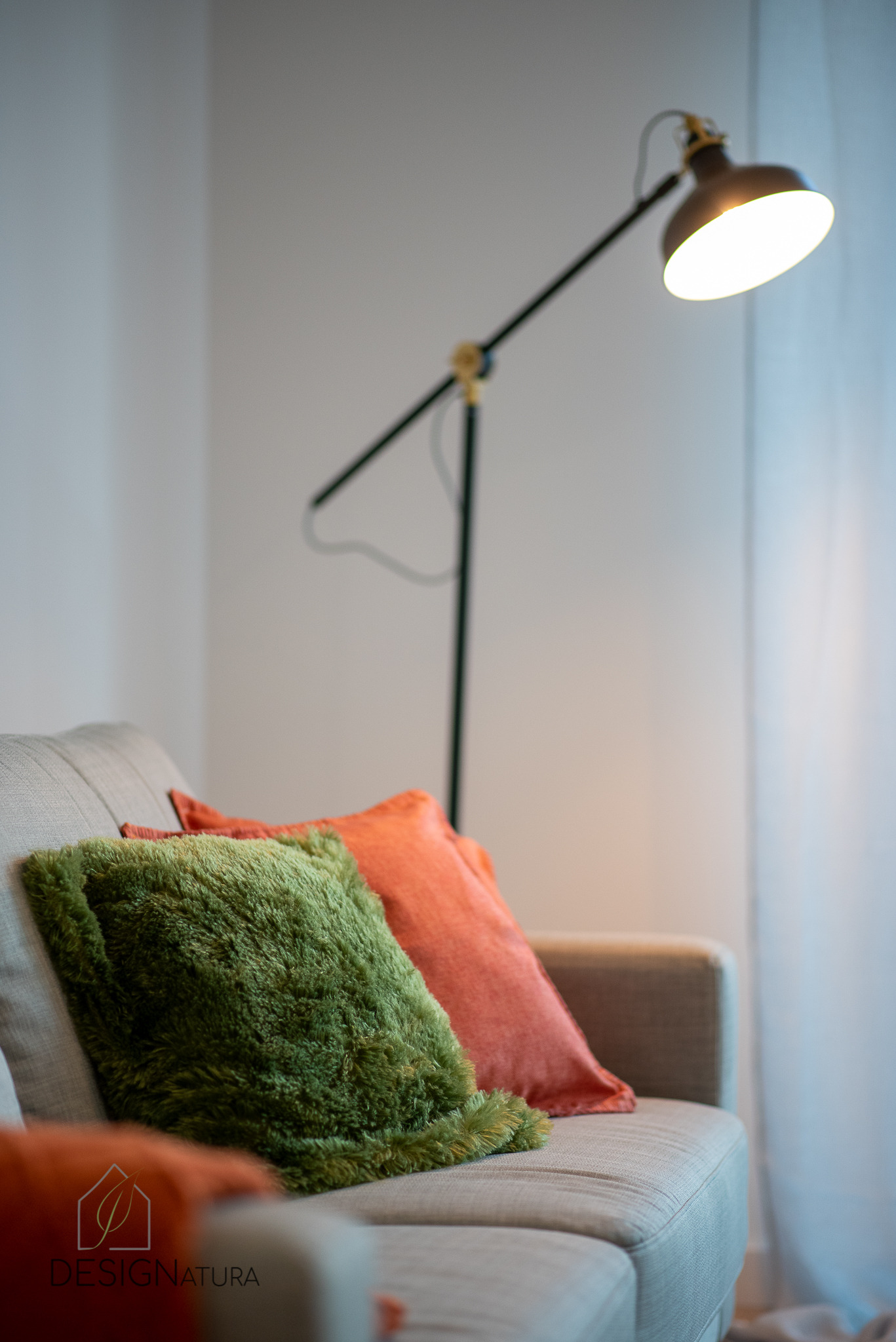 Szary narożnik z dekoracyjnymi poduszkami: zieloną futrzaną i pomarańczową, oświetlony lampą podłogową z regulowanym ramieniem.