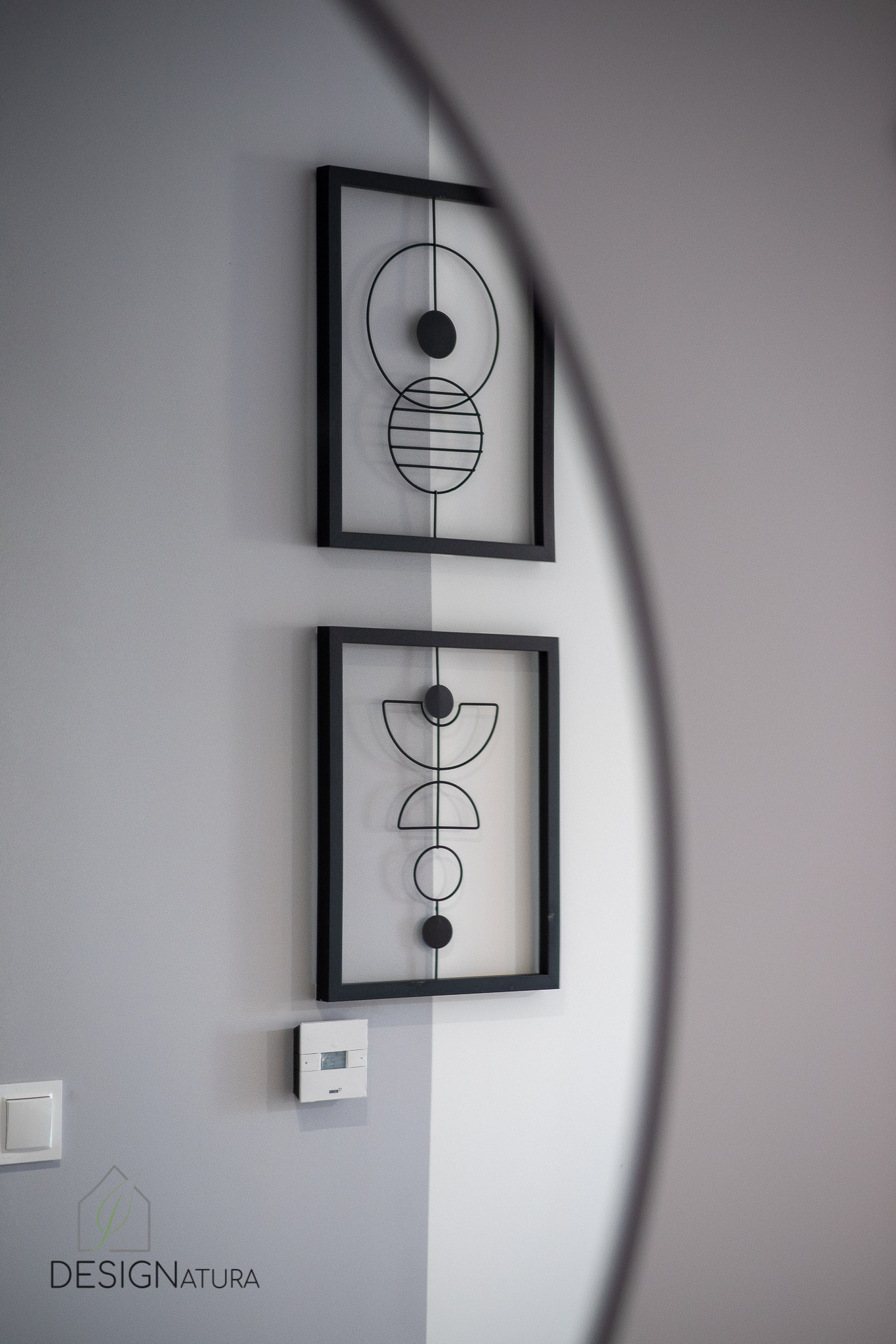 Dwa geometryczne obrazy w czarnych ramkach wiszące na dwukolorowej, szarej ścianie, z widocznym termostatem i włącznikiem światła, częściowo zasłonięte przez element dekoracyjny.