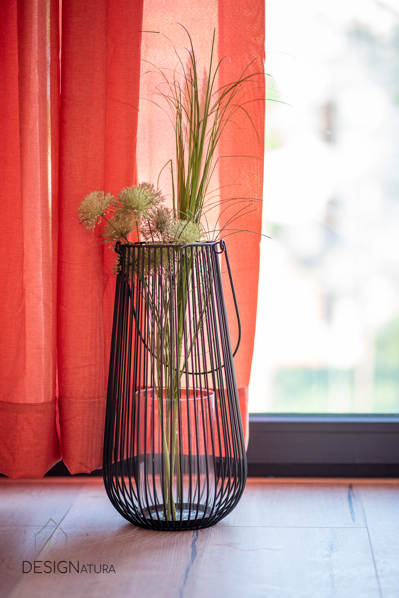 Dekoracyjny, czarny, metalowy lampion z transparentnym wazonem wewnątrz, wypełniony kompozycją z suszonych kwiatów i traw, ustawiony na drewnianej podłodze przy koralowej zasłonie i oknie.
