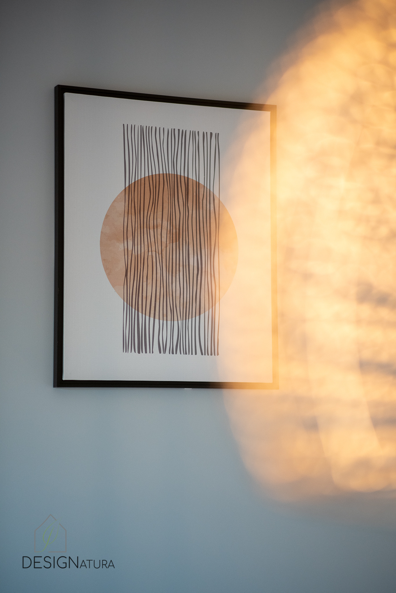 Oprawiony w ramę abstrakcyjny obraz z brązowym kołem i pionowymi liniami na szaro-niebieskiej ścianie, oświetlony ciepłym, rozproszonym światłem słonecznym.