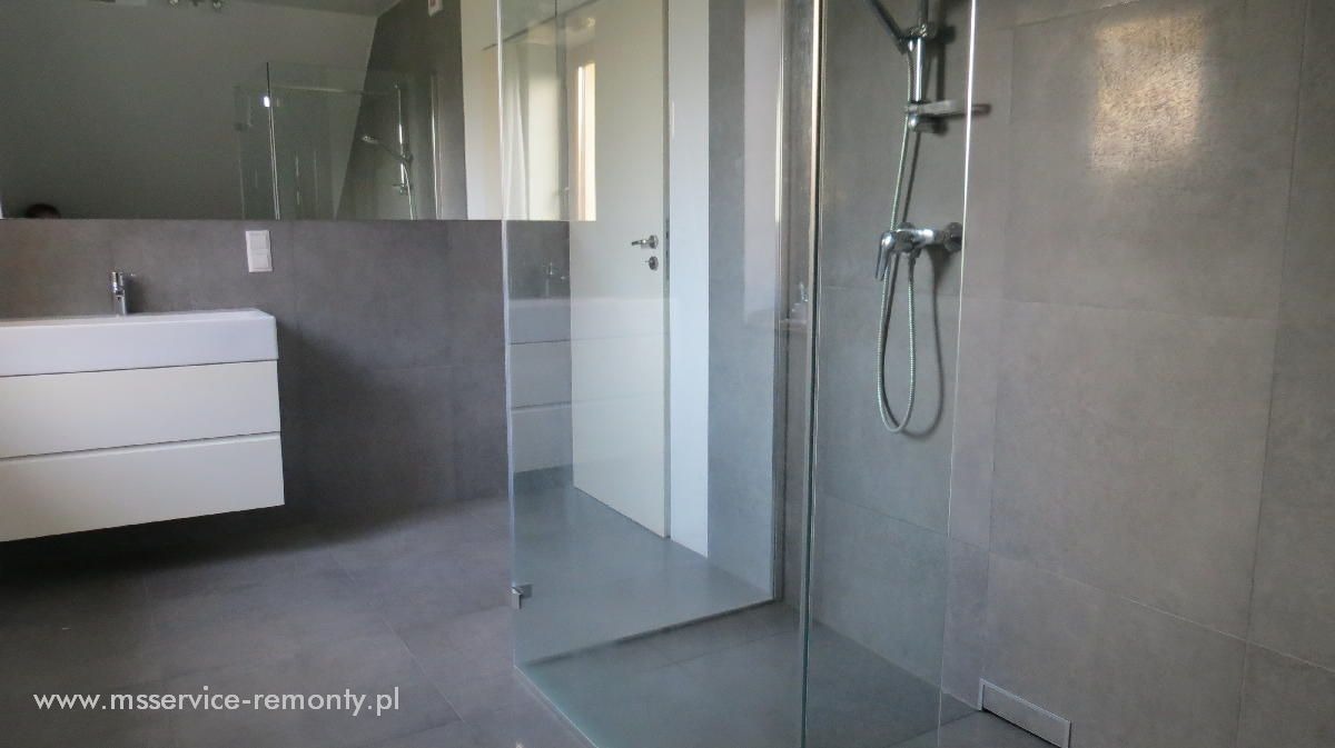 Nowoczesna łazienka z kabiną prysznicową typu walk-in, szare płytki wielkoformatowe, biała szafka pod umywalką z szufladami, minimalistyczna armatura.