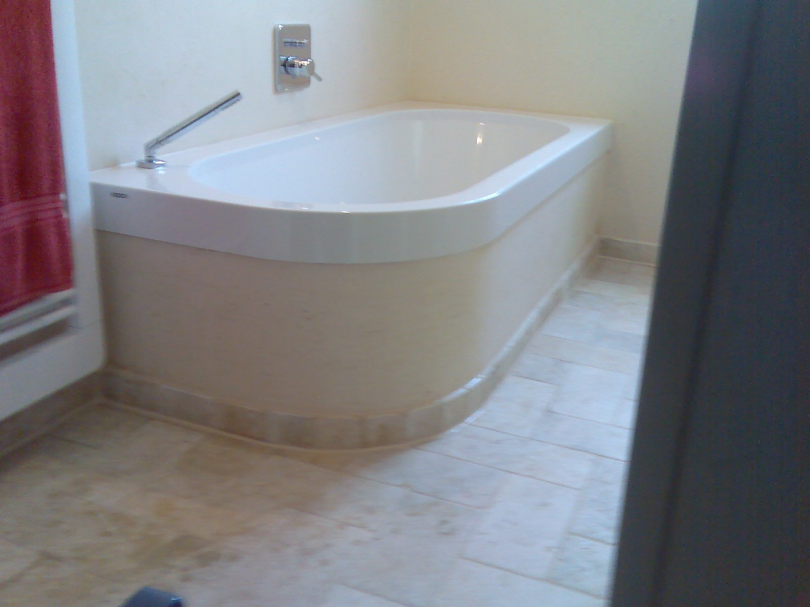 Nowoczesna wanna z obudową w jasnym odcieniu, bateria ścienna, beżowe płytki podłogowe, widok z boku z fragmentem drzwi.