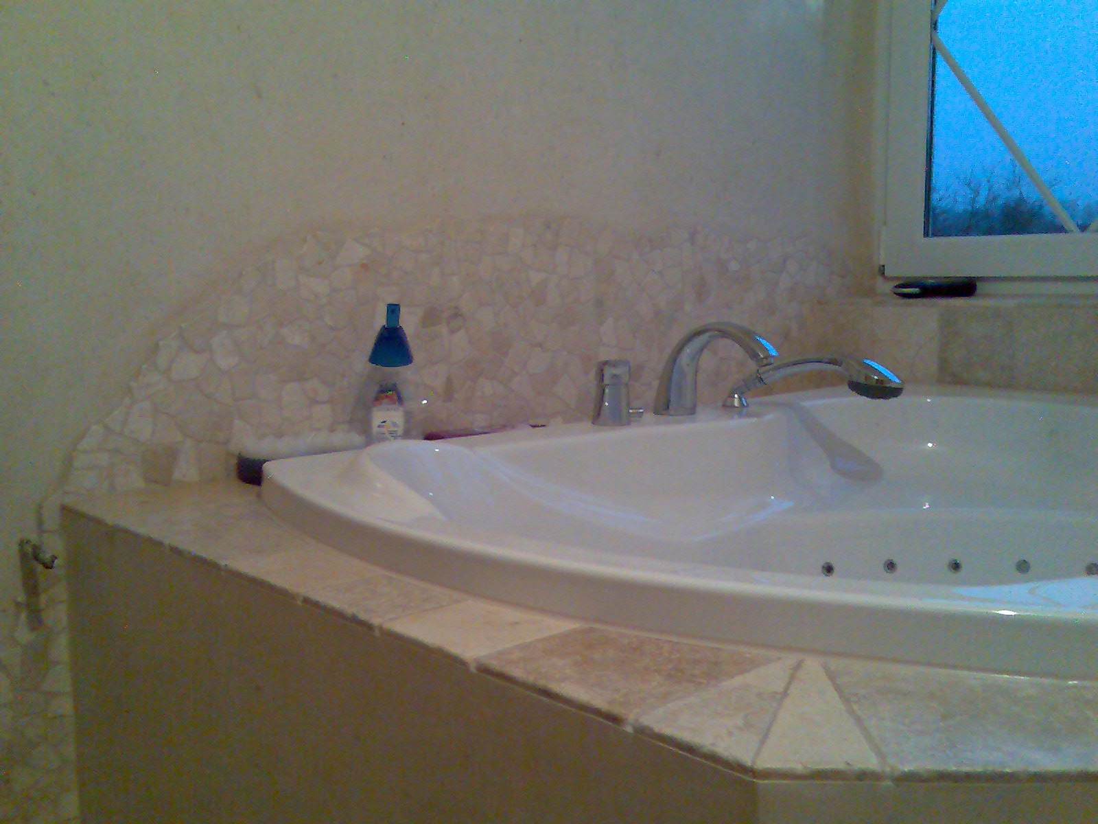 Biała wanna z hydromasażem w narożniku łazienki, z mozaikową ścianą i oknem w tle, widoczna butelka z niebieskim dozownikiem.