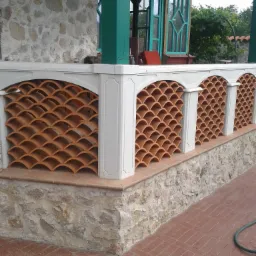 Zadaszony balkon z dekoracyjną balustradą z terakoty w kształcie łusek, białe filary i kamienne wykończenie, widoczne zielone ramy okienne i drzwi.