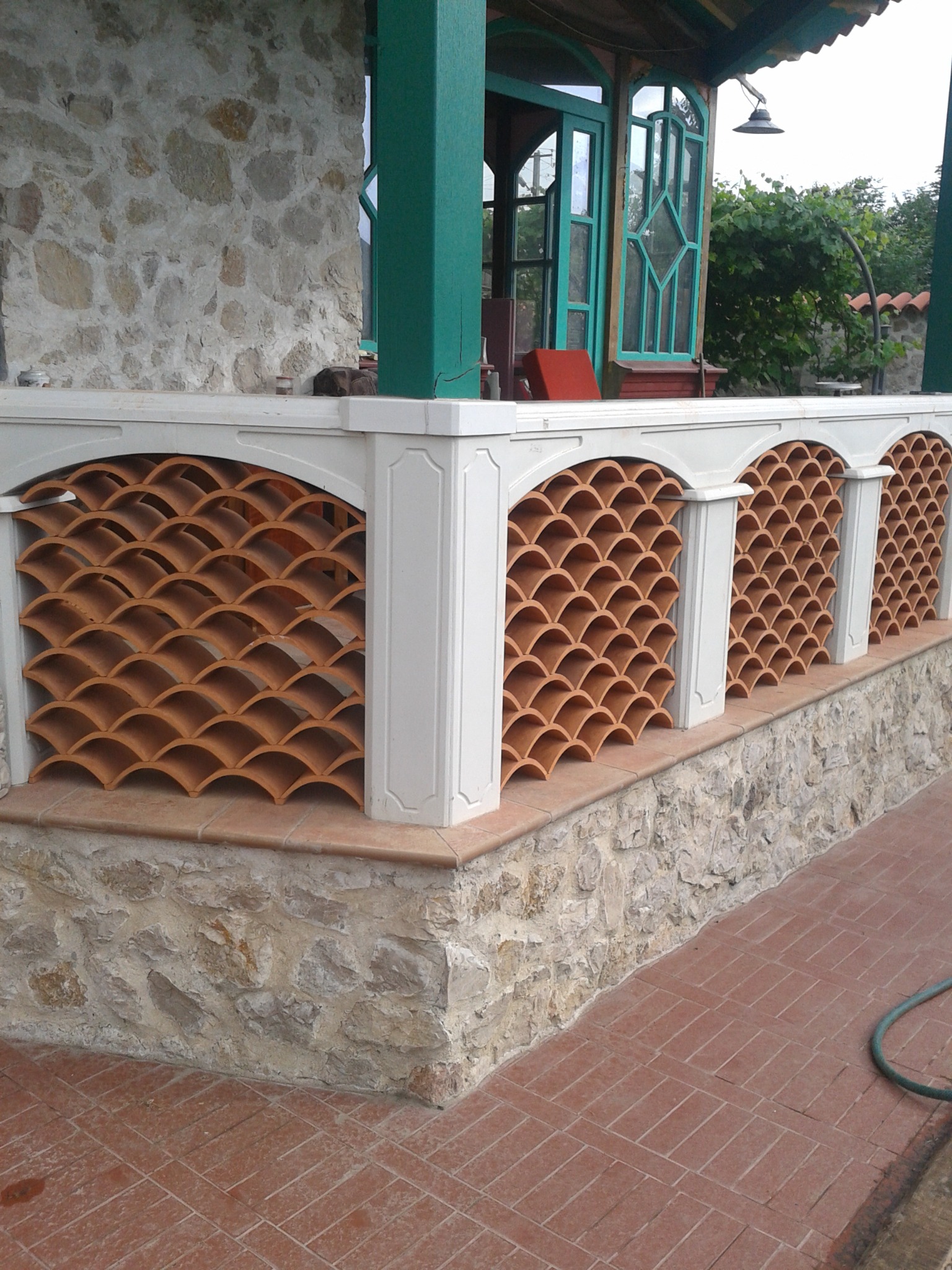 Zadaszony balkon z dekoracyjną balustradą z terakoty w kształcie łusek, białe filary i kamienne wykończenie, widoczne zielone ramy okienne i drzwi.