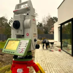 Tachimetr Leica na statywie, na pierwszym planie, w tle pracownicy układający kostkę brukową na posesji z nowoczesnym domem.