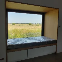 Wnęka okienna z siedziskiem i szafkami w dolnej części, zabezpieczona folią, widok na łąkę za oknem, obok szafka RTV.