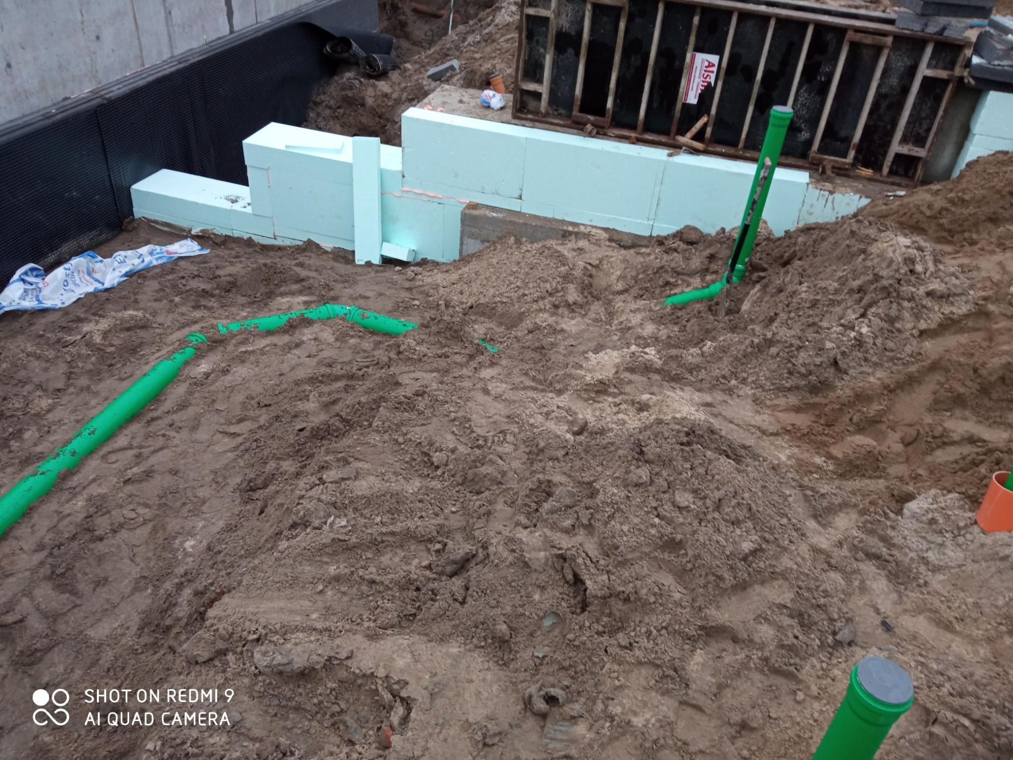 Fundament budynku w trakcie izolacji termicznej styropianem, widoczne wystające rury kanalizacyjne i zasypana ziemia.