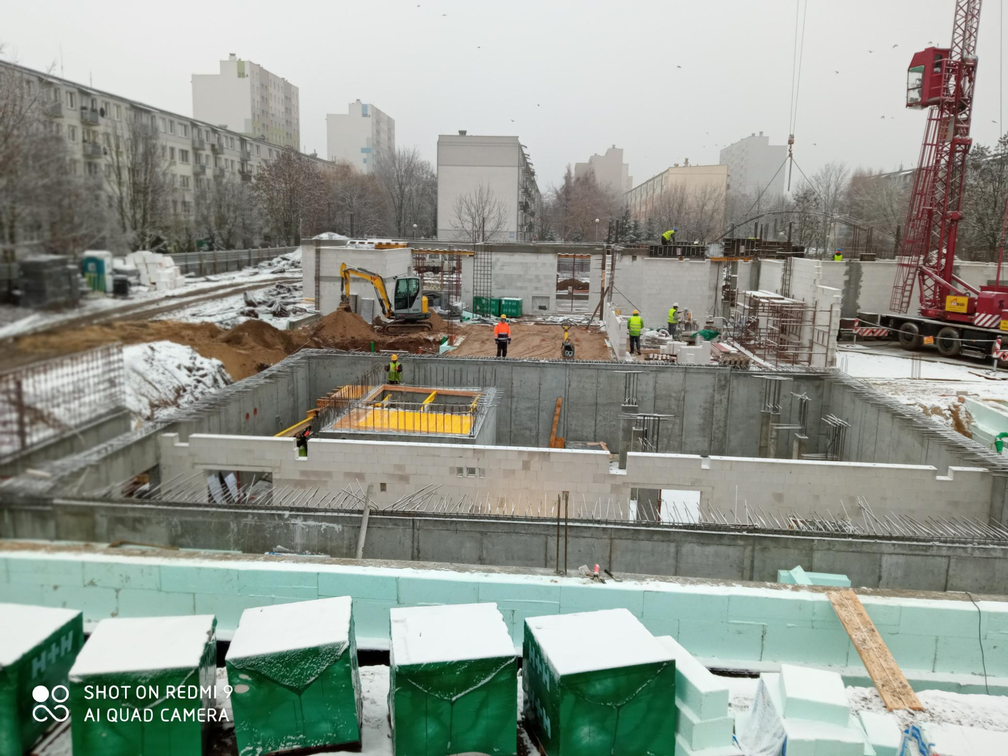 Budowa fundamentów w zimowej scenerii miejskiej, widoczny wykop, zbrojenie, betonowe bloczki i pracujący dźwig