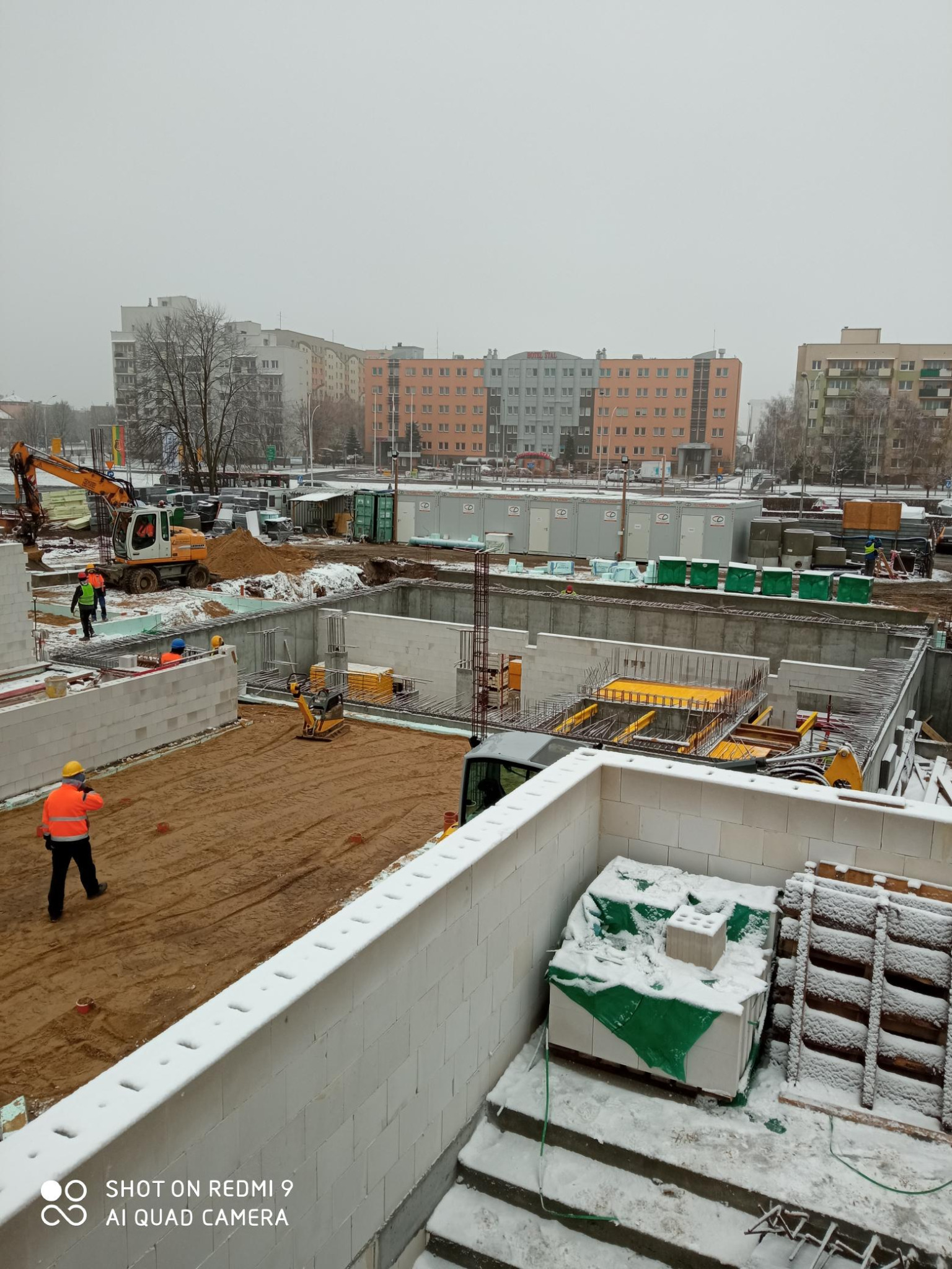Budowa fundamentów budynku mieszkalnego w zimowej scenerii, widoczne zbrojenia, bloczki betonowe, maszyny budowlane i pracownicy w odblaskowych kamizelkach, w tle bloki mieszkalne.