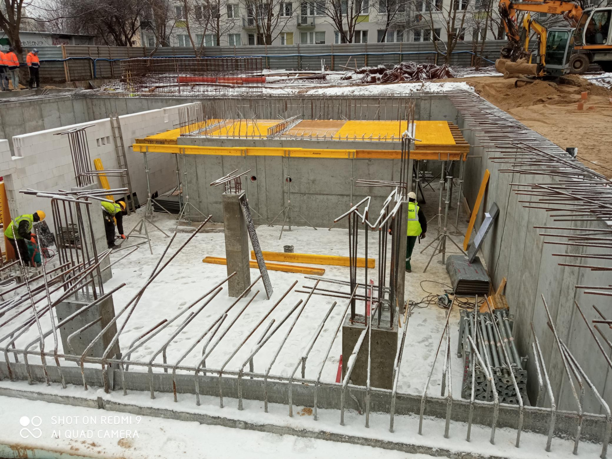 Zimowy plac budowy z wykopem fundamentowym, zbrojeniami pokrytymi śniegiem, żółtym szalunkiem i pracującymi robotnikami w odblaskowych kamizelkach; w tle budynek mieszkalny i koparka.