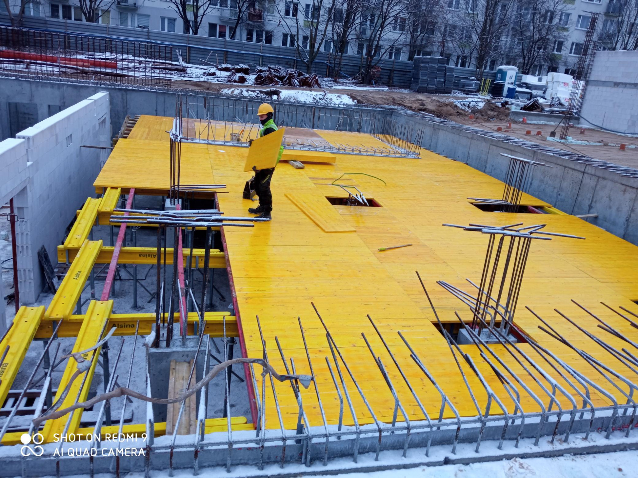 Żółte szalunki na fundamencie budynku w trakcie budowy, widoczny pracownik w kasku i kamizelce odblaskowej przenoszący element szalunku, zima, śnieg na zewnątrz.