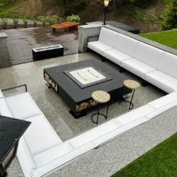 Zewnętrzny salon z białymi sofami modułowymi, czarnym paleniskiem na drewno, dwoma okrągłymi stołkami z mozaiką i oświetleniem ogrodowym, na tarasie z szarej kostki brukowej w deszczowy dzień.