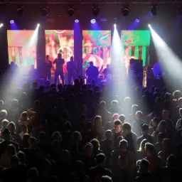 Zespół muzyczny gra na scenie oświetlonej reflektorami przed publicznością w klubie. W tle kolorowe ekrany LED.