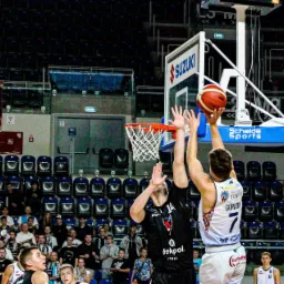 Akcja podczas meczu koszykówki: dwóch zawodników walczy o piłkę przed koszem, kibice na trybunach w tle.