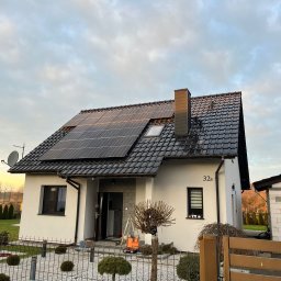 ENERGY INVEST - Biały dom jednorodzinny z ciemną dachówką i panelami słonecznymi na dachu, widoczne drabiny i skrzynka narzędziowa sugerują trwający montaż, ogród z ozdobnymi krzewami i ogrodzeniem.