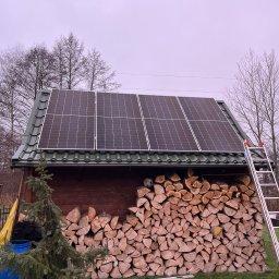 ENERGY INVEST - Mały drewniany budynek z zielonym dachem, na którym zamontowane są panele słoneczne; obok budynku stoi drabina, a pod dachem ułożone jest drewno opałowe, z wystającą piłką do gry.