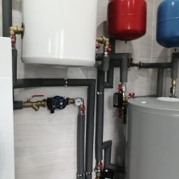 ENERGY INVEST - Instalacja centralnego ogrzewania z białym zbiornikiem z logo, czerwonym i niebieskim naczyniem wzbiorczym, szarym zasobnikiem oraz rurami z izolacją termiczną na tle jasnej, wzorzystej ściany.